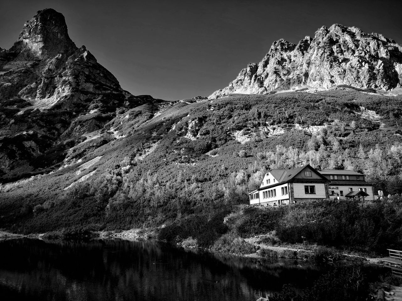 Black and white image of a house by a serene lake with mountains.